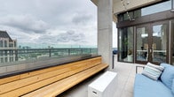 This elevated rooftop terrace offers a striking blend of modern design and urban functionality. The space is defined by its open-air layout, framed by robust concrete columns and floor-to-ceiling glass railings that provide unobstructed panoramic views of the surrounding cityscape and skyline. The light-toned porcelain tile flooring extends throughout, creating a clean and polished aesthetic underfoot. A standout feature is the built-in wooden bench, crafted from warm-toned natural wood planks, which runs along the glass railing perimeter — offering a comfortable, permanent seating area perfectly positioned to take in the sweeping views below. White modular storage units serve a functional purpose while complementing the minimalist design palette. Adjacent to the open terrace, large glass sliding doors connect seamlessly to an interior space, allowing for natural light flow and easy indoor-outdoor access. Track lighting overhead illuminates the transitional covered section, adding ambiance during evening hours. Lush potted greenery near the entryway introduces a touch of nature to the otherwise urban setting. The overall atmosphere is sophisticated, airy, and contemporary, making this terrace an exceptional space for both casual gatherings and quiet moments above the city.