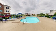 Located at 6312 Clock Tower Dr in Walbridge, Ohio, this community outdoor pool area offers a well-appointed recreational retreat for residents. The centerpiece is a generously sized freeform swimming pool with a smooth, curved design and multiple entry points via stainless steel handrails, making it accessible and inviting. The surrounding concrete sun deck provides ample space for lounging, furnished with a variety of chaise lounges, chairs, and tables shaded by blue patio umbrellas. Manicured landscaping lines the perimeter, adding a touch of greenery that softens the hardscape. The entire area is enclosed by a black wrought iron fence, ensuring safety and privacy while still allowing open sightlines to the broader community and the expansive Ohio sky beyond. An adjacent building with large glass doors provides convenient access to interior amenities. In the background, the community's multi-story brick residential buildings are visible, reflecting traditional architecture with classic detailing. The pool area's open layout and generous deck space make it well-suited for both relaxed individual use and social gatherings, offering a comfortable outdoor amenity in a residential community setting.