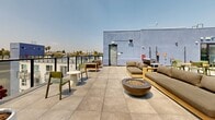Located on Glendale Boulevard in the vibrant Echo Park neighborhood of Los Angeles, this two-level amenity space offers a compelling blend of modern interior design and resort-style outdoor living. The lower level features a stylishly appointed indoor lounge with light wood flooring, soaring double-height ceilings, and bold design accents including a large colorful mural and striking purple accent walls. A sleek kitchenette, comfortable seating arrangements, and a modern staircase with black metal railings complete the interior. Large windows flood the space with natural light and frame views of the surrounding cityscape. The upper level opens to an expansive rooftop terrace that serves as the centerpiece of the space. Here, large-format porcelain tile pavers anchor multiple seating zones furnished with teak-framed sofas, colorful accent chairs, and a fire pit bowl. A dedicated outdoor kitchen with a gas grill and dining table makes the terrace ideal for entertaining. Curated planters with lush greenery add a natural element throughout. Glass panel railings along the perimeter maximize unobstructed panoramic views, including a dramatic east-facing perspective of the Downtown Los Angeles skyline—making this a truly distinctive urban retreat.