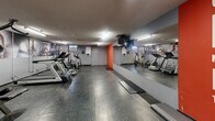 This fitness facility at 75 Charolais Blvd in Brampton, Ontario, is a dedicated exercise room designed to offer a functional and motivating workout environment. The space features durable rubber flooring with a speckled finish, providing both safety and comfort underfoot. Overhead fluorescent lighting ensures the room is well-lit throughout.

The room is equipped with a variety of cardio machines, including treadmills, elliptical trainers, and stationary bikes, as well as a multi-station strength training machine and free weight equipment, catering to a range of fitness needs. A large wall mirror on one side visually expands the space and allows users to monitor their form during workouts.

The interior design incorporates bold motivational graphics, including large-scale athletic photography wrapping the upper walls and a striking orange accent wall featuring inspirational text, contributing to an energetic and encouraging atmosphere. The grey wall tones complement the vivid accents, keeping the space feeling clean and focused.

Access is controlled via a key fob entry system, and informational postings are displayed near the entrance. The overall layout is practical and well-organized, making efficient use of the available floor space.