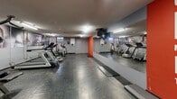 This fitness facility at 75 Charolais Blvd in Brampton, Ontario, is a dedicated exercise room designed to offer a functional and motivating workout environment. The space features durable rubber flooring with a speckled finish, providing both safety and comfort underfoot. Overhead fluorescent lighting ensures the room is well-lit throughout.

The room is equipped with a variety of cardio machines, including treadmills, elliptical trainers, and stationary bikes, as well as a multi-station strength training machine and free weight equipment, catering to a range of fitness needs. A large wall mirror on one side visually expands the space and allows users to monitor their form during workouts.

The interior design incorporates bold motivational graphics, including large-scale athletic photography wrapping the upper walls and a striking orange accent wall featuring inspirational text, contributing to an energetic and encouraging atmosphere. The grey wall tones complement the vivid accents, keeping the space feeling clean and focused.

Access is controlled via a key fob entry system, and informational postings are displayed near the entrance. The overall layout is practical and well-organized, making efficient use of the available floor space.