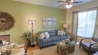 This single-floor space offers a thoughtfully designed living area that balances comfort and style. The living room serves as the focal point, showcasing warm hardwood flooring that adds a natural, inviting feel throughout. A bold sage green accent wall provides a refreshing backdrop, complemented by a triptych landscape artwork that adds artistic character to the space.

The room is furnished with a contemporary tufted gray sofa adorned with decorative throw pillows, creating a comfortable and visually appealing seating arrangement. A rustic wood-and-glass coffee table adds an earthy, organic touch, while an ornate floor lamp with an open-cage design contributes ambient lighting alongside a classic table lamp. A ceiling fan with an integrated light fixture ensures both comfort and illumination.

Natural light filters through patterned curtains, softening the overall ambiance and adding a layer of texture to the room. Lush indoor greenery in a woven basket planter introduces a fresh, natural element that ties seamlessly into the earthy color palette. The overall design aesthetic is warm and cohesive, blending contemporary furnishings with natural materials to create a space that feels both functional and inviting.