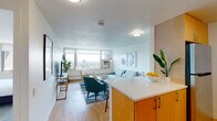 This single-floor residence offers a thoughtfully designed open-concept layout that seamlessly connects the kitchen and living areas. The kitchen is equipped with warm-toned wood cabinetry, stainless steel appliances including a refrigerator, electric range, and microwave, complemented by a light blue subway tile backsplash and a peninsula with a crisp white countertop that serves as both a prep and casual dining surface. Light hardwood flooring runs throughout the main living areas, enhancing the bright, airy atmosphere.

The living room is generously sized, featuring large windows that frame expansive cityscape views and fill the space with abundant natural light. A wall-mounted television and open shelving add functional character to the room.

The bedroom is a calm retreat, finished with plush carpeting and a wide window that continues the sweeping urban panorama. A built-in wardrobe provides ample storage. The bathroom features classic white subway tile surrounding a full soaking tub and shower combination, a floating vanity, and hardwood flooring, offering a clean and timeless aesthetic throughout.