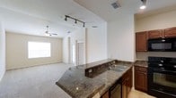 This single-story space showcases an open-concept layout that seamlessly connects the kitchen and living area, creating a spacious and functional environment. The kitchen is anchored by a wraparound dark granite countertop that extends into a peninsula, providing ample prep space and a natural division between the cooking and living zones. A double-basin stainless steel sink is integrated into the countertop, complemented by rich dark wood cabinetry beneath. Stainless steel appliances add a modern touch. The kitchen is illuminated by a track lighting system mounted on the ceiling, offering directed, adjustable light throughout the space. The living area flows naturally from the kitchen, featuring neutral-toned carpet flooring and a ceiling fan that enhances comfort and air circulation. A large window allows natural light to fill the room, contributing to an airy and open atmosphere. The walls are finished in a soft, neutral tone throughout, creating a clean and cohesive aesthetic. A doorway visible in the background suggests additional rooms or access points, adding to the overall functionality of this well-designed, single-level space.