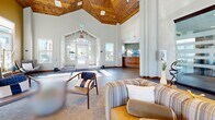 Located in Parker, Colorado, this clubhouse and fitness center is a thoughtfully designed community facility that blends rustic mountain character with modern amenities. The centerpiece of the space is a sweeping great room distinguished by soaring vaulted ceilings with exposed wood beam trusses and warm timber detailing. A floor-to-ceiling stacked stone fireplace anchors the living area, which is bathed in abundant natural light streaming through large windows. Slate-toned tile flooring flows throughout the open common areas, complementing the neutral and earthy color palette. A welcoming reception area greets visitors near the main entrance, featuring a handcrafted wood desk set against sage-green walls. The kitchen and dining area offer a generously sized island with bar seating, stainless steel appliances, warm wood cabinetry, and a tiled backsplash, alongside a bright dining nook with outdoor views. The fitness facilities are divided into two dedicated exercise rooms: one housing cardio and strength equipment including treadmills, ellipticals, and weight benches, and a second studio space equipped with cycling bikes and free weights, both benefiting from large windows and ceiling fans. Connecting hallways provide seamless circulation throughout the facility.