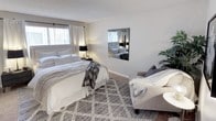 This single-floor space features a generously sized bedroom designed with both comfort and functionality in mind. The room is centered around a king-sized bed dressed in crisp white bedding with layered textures, including a chevron-patterned throw blanket, adding warmth and visual interest. A patterned area rug in neutral tones anchors the space and complements the soft, beige carpeted flooring beneath.

Natural light filters through a window dressed with sheer white curtains, brightening the room's neutral gray and white color scheme. A wall-mounted mirror with a sleek dark frame reflects light throughout the space, enhancing the sense of openness. A bedside table with a black table lamp provides practical ambient lighting.

A cozy seating area is positioned to one side, featuring an upholstered armchair in a warm gray tone, a draped throw blanket, and a small round side table — creating an inviting reading nook. A lush indoor plant adds a natural, refreshing element to the corner. The textured popcorn ceiling is consistent throughout, completing the cohesive, comfortable aesthetic of this well-styled space.