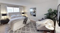 This single-floor space features a generously sized bedroom designed with both comfort and functionality in mind. The room is centered around a king-sized bed dressed in crisp white bedding with layered textures, including a chevron-patterned throw blanket, adding warmth and visual interest. A patterned area rug in neutral tones anchors the space and complements the soft, beige carpeted flooring beneath.

Natural light filters through a window dressed with sheer white curtains, brightening the room's neutral gray and white color scheme. A wall-mounted mirror with a sleek dark frame reflects light throughout the space, enhancing the sense of openness. A bedside table with a black table lamp provides practical ambient lighting.

A cozy seating area is positioned to one side, featuring an upholstered armchair in a warm gray tone, a draped throw blanket, and a small round side table — creating an inviting reading nook. A lush indoor plant adds a natural, refreshing element to the corner. The textured popcorn ceiling is consistent throughout, completing the cohesive, comfortable aesthetic of this well-styled space.