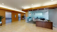 This single-floor commercial lobby presents a well-appointed, professional environment designed to accommodate both functionality and visitor comfort. The space is anchored by warm honey-toned wood wall paneling that frames a pair of stainless steel elevator doors, flanked by tall potted tropical plants that introduce a natural element to the refined interior. Recessed lighting is strategically placed throughout, complementing the cove lighting integrated into the wood beam detailing above.

The reception area features a substantial wood desk positioned adjacent to a frosted glass partition wall, which encloses a semi-private office or administrative zone. A seating chair is visible within this alcove, suggesting a dedicated consultation or waiting area. The main floor is finished in large-format, light travertine-style tiles that extend throughout, creating a sense of openness and continuity.

Two distinct seating arrangements serve visitors: one near floor-to-ceiling windows with sheer roller shades, furnished with sleek black leather armchairs and a glass-top coffee table, and another near the building entrance featuring a matching armchair and loveseat with accent pillows. The generous glazing invites natural light and views of the surrounding outdoor greenery, enhancing the welcoming atmosphere of the lobby.