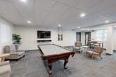 Located at 225 Sandwich St N in Amherstburg, Ontario, this generously sized amenity lounge offers a thoughtfully designed communal space that combines recreation, relaxation, and entertainment in a single, open-plan layout. The room is finished with a neutral-toned carpet featuring a subtle tile pattern, crisp white walls, and a suspended drop ceiling with recessed lighting throughout, creating a clean and well-lit environment.

The games area serves as a central focal point, featuring a full-size billiards table with an elegant dark wood frame, complemented by a wall-mounted cue rack and a flat-screen TV mounted above a floating media shelf. Comfortable accent chairs are positioned nearby, providing seating for spectators. Adjacent to the games area is a relaxed lounge zone with a large sectional sofa, a round wood coffee table, and side tables, all bathed in natural light from vertical blind-covered windows.

The kitchenette area is outfitted with rich dark wood cabinetry, a stainless steel refrigerator, subway tile backsplash, and a spacious granite-topped island with upholstered bar seating — perfect for hosting and casual gatherings. A dining area with bar-height seating rounds out the space. Together, these distinct zones create a versatile and inviting communal retreat.