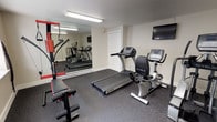 Located at 3000 Austin Drive in Bremerton, Washington, this single-floor fitness space is thoughtfully designed to support a comprehensive workout routine. The room features durable speckled rubber flooring throughout, providing both traction and impact absorption ideal for exercise use.

The space is outfitted with a notable selection of True-brand cardio equipment, including a treadmill, a recumbent stationary bike, and an elliptical machine. A multi-function cable resistance trainer with an adjustable bench serves as the primary strength training station, accommodating a wide variety of exercises. A wall-mounted flat-screen television is positioned for easy viewing during workouts.

A large wall mirror spans one side of the room, visually expanding the space and allowing users to monitor their form. Overhead fluorescent lighting ensures consistent illumination throughout. Windows along the exterior wall allow natural light to enter and offer views of the surrounding outdoor landscape, including a stone feature visible from the exterior. An adjacent doorway connects the fitness room to other areas of the property, contributing to an organized and functional layout.