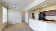 This single-floor, 2-bedroom, 2-bathroom residence features an open-concept layout that connects the kitchen and living area seamlessly. The living room is finished with neutral-toned carpet, crown molding, and track lighting mounted on the ceiling, contributing to a clean, well-lit atmosphere. An arched doorway adds an architectural detail that transitions the space toward the hallway and additional rooms, providing a subtle decorative touch.

The kitchen is positioned along one side of the open layout, defined by a raised breakfast bar counter that overlooks the living area. Dark-stained upper cabinetry contrasts with the light countertops, and a full-size black refrigerator anchors the kitchen space. A pass-through design between the kitchen and living area enhances the sense of openness and allows for easy interaction across both spaces.

Neutral wall tones are consistent throughout, creating a cohesive and versatile interior. Visible HVAC vents indicate a central air system. The hallway, accessible through the arched passage, leads to the bedrooms and bathrooms, keeping the private areas separate from the main living spaces. Overall, the layout is practical and well-organized.