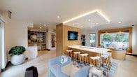 This single-floor community lounge delivers a polished, California-inspired aesthetic through a thoughtfully curated open-plan layout. The space is anchored by a long, white quartz bar counter with warm wood paneling, lined with sculptural white bar stools — an ideal hub for socializing or casual work. Recessed lighting and LED cove lighting along a tray ceiling create a bright, welcoming ambiance throughout.

A dedicated kitchenette sits in one corner, featuring white cabinetry, a stainless steel sink, built-in microwave, and a beverage refrigerator, all framed by a striking geometric blue tile accent wall that serves as a bold visual focal point. Coastal-themed artwork and photography complement the California theme reinforced by a prominent wall-mounted "CALIFORNIA" letter installation flanked by windows that invite natural light and outdoor greenery views.

The lounge seating area offers plush light-blue upholstered chairs and sofas atop a patterned area rug, creating a relaxed gathering zone. Wall-mounted flat-screen televisions are positioned for easy viewing throughout the space. Large windows and a glass-framed opening connect seamlessly to an outdoor patio furnished with additional lounge seating, extending the communal experience outdoors. Light-toned tile flooring and an abundance of indoor plants complete the fresh, airy atmosphere.