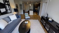 This single-floor residence showcases an open-concept layout that seamlessly connects the living area to the kitchen, creating a spacious and functional flow throughout the home. The living area is anchored by warm hardwood flooring and features a neutral-toned area rug with a geometric pattern, complemented by a sleek, dark round coffee table. A plush blue-gray sofa pairs with a patterned accent chair, contributing to the space's contemporary yet comfortable aesthetic. Track lighting and a statement floor lamp provide layered illumination throughout.

The kitchen is a standout feature, centered around a white island with pendant lighting hanging above, offering both prep space and casual seating. Dark cabinetry on the perimeter contrasts sharply with the lighter island, while stainless steel appliances add a polished, modern touch. A patterned kitchen rug defines the cooking zone and ties the two spaces together visually.

Additional doors visible from the main living area suggest access to a bathroom, closet, and other rooms, all finished with crisp white millwork. The overall ambiance is modern and cohesive, with a balanced contrast of light and dark tones throughout.