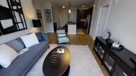 This single-floor residence showcases an open-concept layout that seamlessly connects the living area to the kitchen, creating a spacious and functional flow throughout the home. The living area is anchored by warm hardwood flooring and features a neutral-toned area rug with a geometric pattern, complemented by a sleek, dark round coffee table. A plush blue-gray sofa pairs with a patterned accent chair, contributing to the space's contemporary yet comfortable aesthetic. Track lighting and a statement floor lamp provide layered illumination throughout.

The kitchen is a standout feature, centered around a white island with pendant lighting hanging above, offering both prep space and casual seating. Dark cabinetry on the perimeter contrasts sharply with the lighter island, while stainless steel appliances add a polished, modern touch. A patterned kitchen rug defines the cooking zone and ties the two spaces together visually.

Additional doors visible from the main living area suggest access to a bathroom, closet, and other rooms, all finished with crisp white millwork. The overall ambiance is modern and cohesive, with a balanced contrast of light and dark tones throughout.