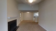 This single-floor space is designed with an open-concept layout that connects the main living area to the kitchen, creating a sense of spaciousness and ease of movement. The living room is a notable focal point, anchored by a tile-surround fireplace with a clean white mantle that adds a classic touch to the neutral interior.

The ceilings rise higher than standard, with a stepped architectural detail that adds visual depth to the space. A pendant light fixture hangs centrally, providing ambient illumination to the carpeted living area, which features wall-to-wall neutral-toned carpet throughout.

The kitchen is visible from the main living space and is equipped with white cabinetry, a range, and under-cabinet lighting. A pass-through counter or peninsula connects it to the main area, enhancing the open flow. A hallway to the side suggests access to additional rooms beyond the main living space. The overall color palette is consistent and neutral, with beige walls and white trim providing a clean, move-in-ready aesthetic throughout.