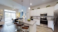 This single-floor residence showcases a thoughtfully designed open-concept layout that seamlessly connects the kitchen and living areas. The kitchen is a standout feature, boasting crisp white cabinetry that extends to impressive heights, a classic white subway tile backsplash, and a full suite of stainless steel appliances including a range, microwave, and dishwasher. At the heart of the kitchen sits a generously sized island topped with light granite countertops, illuminated by stylish pendant lights and lined with modern bar stools — perfect for casual dining or entertaining. The open living area beyond benefits from soaring vaulted ceilings and abundant natural light streaming through large floor-to-ceiling windows dressed with vertical blinds. Light wood-look flooring runs throughout, adding warmth and continuity between spaces. A ceiling fan in the living area enhances comfort, while colorful artwork and carefully placed greenery on top of the upper cabinets lend a vibrant, curated aesthetic. The overall ambiance is bright, airy, and contemporary, offering a welcoming and functional environment that balances modern finishes with an inviting sense of openness.