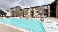 Located in Katy, Texas, this amenity space is part of a contemporary multi-story residential community defined by modern architecture featuring clean lines, neutral tones, and private balconies. The expansive outdoor pool area serves as a central focal point, with a generously sized, sparkling turquoise pool surrounded by lounge chairs and manicured palm trees, evoking a resort-like atmosphere. The pool deck provides ample space for relaxation against the backdrop of the community's striking façade.

The fitness center is a well-appointed, spacious facility designed to accommodate a full range of workout routines. Large windows along one wall bring in natural light and offer views of the pool area, creating an open, energizing environment. The flooring combines light hardwood with teal-toned exercise mats, adding a vibrant, modern aesthetic throughout. Cardio equipment, including treadmills, ellipticals, and stair climbers, lines the perimeter, while a dedicated free weights section and functional training stations—including cable machines and punching bags—provide extensive strength training options. A mirrored studio room with glass-paneled French doors offers space for yoga, stretching, or group fitness activities. Recessed and tray ceiling lighting completes the polished, professional atmosphere.