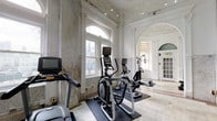This fitness center occupies a strikingly elegant historic space, blending classical architectural grandeur with modern exercise functionality. The interior is defined by soaring ceilings adorned with intricate plasterwork and ornate cornice detailing, creating a dramatic sense of vertical space rarely found in a gym setting. Marble wainscoting lines the walls throughout, complemented by marble tile flooring that reinforces the refined, heritage character of the space.

Tall, arched windows along the exterior walls flood the interior with abundant natural light, offering street-level views while enhancing the open, airy ambiance. Stately white columns with decorative capitals divide the space into distinct zones, adding architectural rhythm and depth. Arched doorways connect the main exercise area to an adjoining room, maintaining visual continuity across the layout.

The main workout area is equipped with cardio machines including treadmills, ellipticals, and a rowing machine, all positioned on protective floor mats. A dedicated free weights zone features a full dumbbell rack and weight bench, set against paneled white walls beneath motivational fitness artwork. Glass-paneled entry doors and string lighting overhead add a contemporary touch, while the overall atmosphere strikes a compelling balance between historic elegance and practical fitness functionality.