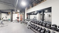Located at 4901 Wornall Rd in Kansas City, Missouri, this fitness center offers a comprehensive workout facility designed to accommodate a wide range of exercise needs. The space features an industrial aesthetic, with exposed ceiling infrastructure, dark-painted overhead pipes and ducts, and concrete masonry block walls that lend a raw, functional character to the environment.

The gym is divided into distinct workout zones. The free weights area is a standout feature, housing an extensive rack of dumbbells spanning a wide range of weights, positioned alongside full-length wall mirrors that enhance the sense of space and allow users to monitor their form. Adjacent to this area, a power rack setup with barbells and weight plates supports strength training and compound lifts.

The cardio section is well-appointed with multiple machines, including ellipticals, a stationary bike, and a rowing machine, all arranged to allow comfortable use. Additional functional training equipment includes battle ropes, a captain's chair, and a weight bench, supporting varied training styles.

Durable rubber flooring covers the entire area, providing cushioning and traction throughout. Recessed and overhead lighting ensures the space is well-lit. A wall-mounted television and a sliding barn-style door add practical and aesthetic touches. The layout is efficient and organized, making this fitness center a fully equipped facility for strength, cardio, and functional training.