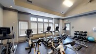 Located on Rio Grande Court in Saint Charles, Missouri, this fitness center offers a comprehensive workout environment designed to support a wide range of exercise routines. The space features a striking vaulted ceiling with a ceiling fan, giving the room an open, airy feel that is enhanced by large windows along the walls that flood the interior with natural light. The flooring throughout is a rich, dark hardwood-style material that adds a polished, modern aesthetic.

The cardio zone is well-appointed with multiple treadmills, upright and recumbent stationary bikes, spinning cycles, and elliptical machines, all from quality brands. A dedicated strength training area includes a multi-station cable and weight machine, a free weight rack stocked with a full range of dumbbells, medicine balls, and a stability ball, along with a weight bench for targeted exercises. Full-length mirrors line one wall, creating a sense of expanded space while allowing users to monitor their form. Multiple flat-screen televisions are mounted from the ceiling, keeping members entertained during workouts.

A hand sanitizer station is conveniently positioned near the entrance. The overall ambiance is clean, bright, and professionally maintained, making this fitness center a functional and inviting space for residents seeking a convenient on-site workout facility.