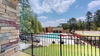 Located at 3130 Hillandale Rd in Durham, NC, this outdoor amenity space is centered around a generously sized resort-style swimming pool with crystal-clear blue water. The pool deck is outfitted with an extensive row of lounge chairs featuring bold red cushions, providing ample space for sunbathing and relaxation. Built-in in-pool loungers offer a unique way to cool off while soaking up the sun. The area is enclosed by a classic black iron fence, lending a sense of security while maintaining open, scenic views of the surrounding mature pine and hardwood trees that create a lush, natural backdrop. A well-appointed clubhouse or pool house anchors one end of the deck, constructed with warm tan horizontal siding, stone accents, and a covered porch supported by distinctive stacked-stone columns. The covered porch is furnished with comfortable wicker lounge seating and provides direct access to the interior through French-style doors. Additional outdoor seating areas and shaded spots with red umbrellas are dispersed throughout the deck. The neighboring multi-story residential building is visible in the background, framed by the verdant wooded setting. The overall ambiance is bright, welcoming, and resort-like, ideal for outdoor recreation and leisure.