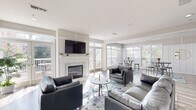 This well-appointed clubhouse in Owings Mills, MD offers a thoughtfully designed single-floor layout that balances comfort and functionality. The living room serves as the heart of the space, featuring a stone-accented fireplace topped with a wall-mounted flat-screen TV, elegant wainscoting panels, and rich hardwood-style flooring. Plush leather seating arranged around a glass coffee table creates a welcoming gathering area, while abundant natural light pours in through large windows and a set of French doors that open onto an outdoor deck with views of the surrounding greenery. The dining area is bathed in natural light from floor-to-ceiling windows, furnished with high-top bistro tables and bar stools, making it ideal for casual dining or social gatherings. A pass-through window connects the dining space seamlessly to the kitchen, which is outfitted with crisp white shaker cabinetry, granite countertops, stainless steel appliances, and a double sink. The cohesive neutral color palette, colorful area rugs, and consistent hardwood-style flooring throughout lend the entire clubhouse a clean, modern aesthetic that is both inviting and versatile.