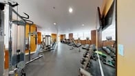 Located at 1630 Aliceanna Street in Baltimore's vibrant Canton neighborhood, this well-appointed fitness center offers a comprehensive workout environment within a single, open-plan floor. The space is defined by its bold orange accent walls, exposed brick detailing, and rubber-floored workout areas, creating an energetic yet functional atmosphere.

The cardio zone lines the brick-accented wall and features a row of TRUE-brand treadmills, elliptical trainers, and a stationary cycle, each equipped with individual display screens. Large windows along this wall allow natural light to fill the space, contributing to an open, airy feel during workouts.

The strength training area is equally well-equipped, showcasing a full dumbbell rack with an extensive weight range, cable and multi-station weight machines, adjustable benches, and free-weight accessories including medicine balls and exercise mats. A wall-mounted flat-screen television is positioned for visibility across the floor.

Recessed ceiling lighting supplements the natural light from the multiple windows, ensuring the space remains bright and inviting at all times. The rubber flooring throughout provides both durability and safety underfoot. A large mirror on one wall enhances the sense of space and supports form-focused training. The overall design balances industrial character—through its brick walls and column details—with modern fitness functionality.