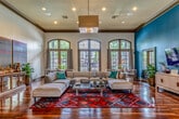 This single-floor office space combines elegant design with a highly functional layout, creating a professional environment that feels both welcoming and refined. Throughout the space, rich hardwood flooring in a deep mahogany tone serves as a unifying element, complemented by soaring ceilings adorned with crown molding and recessed lighting. Bold teal accent walls lend a distinctive character to the interior, contrasting beautifully with crisp white walls and dark wood trim.

The main lounge area is generously proportioned, featuring a large sectional sofa arrangement anchored by a vibrant geometric area rug, a wall-mounted flat-screen TV, and a media credenza flanked by lush potted plants. A series of arched, multi-pane windows along one wall allows abundant natural light to pour in while offering views of the surrounding exterior.

The reception and workstation area provides a practical yet stylish welcome space, equipped with multiple desks, computer workstations, and seating for visitors. Arched French doors and tall transom windows maintain the architectural continuity of the space.

A private office area features a full-size executive desk, a large decorative mirror, and vibrant triptych artwork on the wall. A cozy consultation nook with a round wooden table and upholstered chairs sits beside floor-to-ceiling windows, ideal for intimate meetings. Together, these areas create a cohesive, sophisticated workspace.