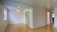 This single-floor space offers a straightforward and functional layout designed for comfortable everyday living. The main living area is characterized by warm, light-toned hardwood flooring that extends throughout, adding a sense of warmth and continuity to the space. Crisp white walls create a bright, airy atmosphere that reflects natural and artificial light effectively throughout the room. A ceiling fan is centrally mounted, providing both ventilation and a subtle decorative touch to the living area. An open rectangular archway connects the main room to an adjacent hallway, offering a natural transition between spaces. The hallway beyond leads to additional areas of the unit and features a fire safety device mounted on the wall, indicating attention to safety standards. The clean baseboards and neutral finishes throughout provide a versatile backdrop suitable for a variety of interior styles and furnishing choices. The overall design is simple and uncluttered, emphasizing functionality and ease of use. The consistent flooring and cohesive color palette across rooms contribute to a unified and well-maintained appearance throughout this single-level space.