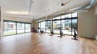 This single-floor fitness studio is thoughtfully designed to deliver a premium workout environment that blends modern aesthetics with functional performance space. The spin and cycling area is the centerpiece of the space, featuring multiple stationary bikes arranged across a generous expanse of warm, richly toned hardwood flooring that runs throughout. Floor-to-ceiling black-framed glass walls flood the interior with natural light and offer unobstructed views of a landscaped outdoor courtyard, creating an energizing and connected atmosphere.

Ceiling fans are strategically placed overhead to ensure consistent airflow, while exposed ductwork adds an industrial-modern character to the otherwise clean, neutral-toned interior. The open layout allows for flexible use of the floor space beyond cycling, accommodating stretching, yoga, or bodyweight training.

A built-in media and storage wall serves as a stylish focal point, featuring a wall-mounted display framed in decorative mosaic tile, flanked by wine rack-style cubbies and lower open shelving for towels and accessories. Dark-framed glass partition doors connect this area to an adjacent fitness room, maintaining visual openness while defining distinct zones. Large mirrors on one wall further enhance the sense of spaciousness. Together, these elements create a cohesive, well-appointed studio that is both functional and visually striking.