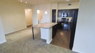 This single-floor residence offers a well-designed open-concept layout that seamlessly connects the kitchen and living areas. The kitchen is a standout feature, showcasing rich dark cabinetry that provides ample storage alongside a suite of black appliances, including a range, microwave, and refrigerator. A central island with a granite-style stone countertop serves as a natural focal point, offering additional prep space and casual seating with bar-height bar stools. Pendant lighting hangs above the island, adding a warm, focused ambiance to the cooking and dining area. The kitchen transitions to hardwood-style flooring, while the adjoining living space is finished with plush carpet, creating a comfortable and distinct separation between zones. The open layout allows natural sightlines throughout, contributing to an airy and spacious feel. A dedicated in-unit laundry area is conveniently accessible just off the main living space, adding a practical touch to everyday living. Track lighting in the living area provides flexible illumination, complementing the overall warm and neutral color palette that gives the space a welcoming, move-in-ready character.