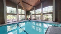 This indoor pool facility is defined by its impressive architectural design, combining natural materials with an abundance of natural light to create a serene, resort-inspired environment. The centerpiece is a generously sized rectangular swimming pool with clear, blue-tinted water, finished with tiled edges and a smooth surrounding deck area.

The structure is distinguished by its soaring vaulted ceiling, constructed from warm-toned wood planks supported by exposed timber beams and posts. This design element adds considerable height to the space, enhancing its open and airy character. Surrounding the pool on multiple sides are expansive floor-to-ceiling windows that frame lush, mature greenery and wooded views beyond a fenced perimeter, seamlessly connecting the interior to the natural landscape outside.

Natural light pours through the large glazed panels, reflecting off the pool's surface and contributing to the bright, welcoming atmosphere. A poolside deck area provides space for seating, with a small number of chairs positioned near the windows. The combination of natural wood tones, ample sunlight, and verdant outdoor views gives the facility a tranquil, nature-integrated ambiance well-suited for year-round use.