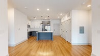 This thoughtfully designed single-floor residence offers a seamless blend of modern style and functional living. The heart of the home is an open-concept kitchen and living area, where warm hardwood-style flooring flows throughout. The kitchen is a standout, featuring a striking two-tone cabinetry scheme — white upper cabinets paired with slate-blue lower cabinets — complemented by a classic white subway tile backsplash and stainless steel appliances including a refrigerator, range, and microwave. A centrally positioned island finished in a coordinating blue-gray tone provides additional workspace and sink access, topped with crisp white countertops and illuminated by a pair of industrial-style pendant lights. The open living area is bright and airy, with recessed lighting overhead and access to natural light through large windows and a glass-paned exterior door. Bedrooms are carpeted for comfort, finished with clean white walls that create a calm, neutral atmosphere. The primary suite includes access to a spacious walk-in closet outfitted with wire shelving on three walls. The primary bathroom is well-appointed with a long dual-sink white vanity, a large wall mirror, black hardware accents, hardwood-style flooring, and a glass-enclosed shower, offering a clean and contemporary aesthetic throughout.