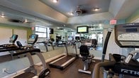 This fitness center is a dedicated, single-floor workout facility designed to support a comprehensive cardio training experience. The room is outfitted with a selection of True-brand cardio equipment, including treadmills, recumbent bikes, and elliptical machines, all arranged to maximize space and usability. Floor-to-ceiling mirrors line the walls, visually expanding the room and allowing users to monitor their form during exercise. A wall-mounted flat-screen television is positioned at eye level, providing entertainment during workouts. Ceiling fans are installed throughout, promoting air circulation and comfort during intense sessions. The flooring consists of durable, dark rubberized material well-suited for a high-traffic fitness environment. Recessed lighting and can lights provide consistent, even illumination throughout the space. Natural light filters in through windows, contributing to an open and energizing atmosphere. The tray ceiling adds an architectural touch that elevates the overall aesthetic of the room. The layout is thoughtfully organized, offering clear pathways between machines and an uncluttered environment conducive to focused, effective exercise sessions.