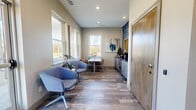 This single-floor hallway space serves as a functional and stylish transitional area, thoughtfully designed to create a welcoming first impression. The layout flows from a bright, glass-door entry corridor into a more expansive lounge and meeting nook, offering a versatile environment suited for reception or small gatherings.

The hallway is characterized by light wood-look plank flooring that runs throughout, complemented by neutral warm-gray walls and recessed lighting overhead. A striking blue textured accent wall draws the eye, adding depth and a contemporary design element. Wooden doors with modern hardware line the corridor, leading to additional rooms including what appears to be a fitness area.

The lounge section transitions naturally into a sitting area furnished with plush blue swivel chairs positioned near large double-hung windows that flood the space with abundant natural light and offer pleasant outdoor views of a landscaped setting. Vaulted ceilings in this area enhance the sense of openness.

A compact conference nook features a clean white rectangular table with coordinating blue chairs, accompanied by a wall-mounted abstract painting and a low-profile storage credenza with cabinetry. The combination of functional furnishings, curated artwork, and thoughtful material choices creates a polished yet inviting ambiance throughout the space.