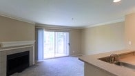 This single-floor residence offers a comfortable and functional layout highlighted by several notable architectural details. The main living area is finished with wall-to-wall carpeting and features crown molding along the ceiling perimeter, lending a clean, polished look to the space. A fireplace with a classic white mantel anchors one wall, serving as a natural focal point in the room.

Natural light fills the living area through large sliding glass doors, which open to a balcony and provide an outdoor view of surrounding greenery. Sheer vertical blinds frame the doors, allowing for adjustable light control while maintaining an airy feel. The neutral beige wall tones complement the light-colored carpet and contribute to a warm, understated ambiance throughout.

Adjacent to the living area, a peninsula counter with a granite-style surface marks the transition to the kitchen space. This open-concept boundary allows for easy interaction between the two areas. The kitchen counter provides both practical workspace and informal seating potential. Overall, the space presents a straightforward, well-maintained layout with classic finishes suitable for comfortable everyday living.