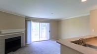 This single-floor residence offers a comfortable and functional layout highlighted by several notable architectural details. The main living area is finished with wall-to-wall carpeting and features crown molding along the ceiling perimeter, lending a clean, polished look to the space. A fireplace with a classic white mantel anchors one wall, serving as a natural focal point in the room.

Natural light fills the living area through large sliding glass doors, which open to a balcony and provide an outdoor view of surrounding greenery. Sheer vertical blinds frame the doors, allowing for adjustable light control while maintaining an airy feel. The neutral beige wall tones complement the light-colored carpet and contribute to a warm, understated ambiance throughout.

Adjacent to the living area, a peninsula counter with a granite-style surface marks the transition to the kitchen space. This open-concept boundary allows for easy interaction between the two areas. The kitchen counter provides both practical workspace and informal seating potential. Overall, the space presents a straightforward, well-maintained layout with classic finishes suitable for comfortable everyday living.