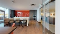 Located at 25 Lewis Street in Boston's East Boston neighborhood, this single-floor amenity space offers a thoughtfully designed environment that blends modern aesthetics with functional versatility. Warm-toned hardwood flooring flows throughout, providing a cohesive foundation that unifies the various areas of the space.

The lounge area invites relaxed gathering with contemporary sectional seating, designer accent chairs, and a curated area rug layered over rich hardwood floors. Floor-to-ceiling windows flood the space with natural light and frame striking waterfront views of Boston Harbor, creating an open and airy ambiance.

A formal conference room anchors the space with a substantial wood-topped table surrounded by vibrant red-orange upholstered chairs. A dramatic starburst pendant light fixture illuminates the room, while expansive glass doors open directly to an outdoor terrace overlooking the harbor — ideal for meetings or private events.

The gourmet kitchen is a standout feature, showcasing a bold orange glass backsplash, a large white island with bar seating, dark wood cabinetry, stainless steel double wall ovens, and sleek glass-panel sliding doors. The kitchen is well-appointed for catering and entertaining at scale.

Throughout the space, glass partitions and transparent architectural elements maintain visual connectivity between zones while offering flexibility and privacy where needed.