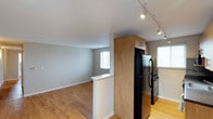 This single-floor space showcases an open-concept layout that seamlessly connects the kitchen and living area, creating a functional and cohesive environment. The kitchen is equipped with black appliances, including a full-size refrigerator and range, complemented by warm wood-tone upper cabinetry that adds a natural contrast to the neutral gray walls. A partial dividing wall with a white countertop serves as a subtle boundary between the kitchen and the adjoining living area, maintaining an open and airy feel without fully separating the two spaces. Track lighting runs along the kitchen ceiling, providing adjustable, directed illumination throughout the area. The living space benefits from multiple windows dressed with white blinds, allowing natural light to filter in and brighten the interior. Light-toned hardwood-style flooring extends throughout both areas, tying the space together cohesively and contributing to the overall clean, contemporary aesthetic. The white baseboards and smooth ceilings further reinforce the space's modern, well-maintained character, making it a practical and visually consistent living environment.