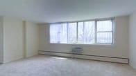 This single-floor space is designed with simplicity and functionality in mind. The main living area is immediately welcoming, characterized by an abundance of natural light streaming through a wide bank of windows that span nearly the full length of one wall. Fitted with vertical blinds, these windows offer flexible light control while framing pleasant outdoor views of mature trees. The neutral, light-toned walls contribute to the bright and airy atmosphere of the room. Underfoot, plush wall-to-wall carpeting in a soft gray hue adds warmth and comfort to the space. Climate control is well-considered, with a baseboard heating system running along the base of the windowed wall, supplemented by a window-mounted air conditioning unit, ensuring year-round comfort. The clean, uncluttered layout of the room allows for a variety of furniture arrangements, making it a versatile space adaptable to different needs. The overall ambiance is calm and understated, with a functional design that prioritizes natural light and a comfortable living environment.