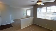 This single-floor space presents a clean, functional layout designed for comfortable everyday living. The interior is finished in soft, neutral gray tones throughout, creating a calm and cohesive ambiance that makes the space feel bright and welcoming.

The main living area is defined by plush wall-to-wall carpeting in a warm beige hue, which extends seamlessly across the open floor plan. A low half-wall partition gracefully divides the space, offering a subtle separation between areas while maintaining an open and airy feel. This architectural detail adds structure to the layout without compromising the sense of spaciousness.

Natural light plays a significant role in the atmosphere of this space. Multiple windows fitted with horizontal blinds allow for adjustable light control, and the views of surrounding greenery further enhance the connection to the outdoors. The windows are positioned to draw daylight deep into the interior, keeping the space feeling open throughout the day.

A ceiling fan with a light fixture is installed overhead, contributing both to air circulation and ambient lighting. The walls are finished with clean white baseboards and trim, providing a crisp contrast to the gray walls.

Overall, this space offers a straightforward, well-maintained environment with thoughtful design details that balance practicality and comfort in a simple, inviting single-floor layout.