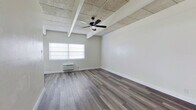 This efficient one-bedroom, one-bathroom unit is thoughtfully laid out across a single floor, making the most of its compact footprint. The main living area features durable grey wood-style plank flooring that runs throughout, crisp white walls, and a distinctive ceiling with exposed beams and textured panels that add a subtle industrial character. A ceiling fan with integrated lighting anchors the space overhead, providing both air circulation and ambient light. A wall-mounted HVAC unit and a window offering natural light complete the living area.

The kitchen is clean and functional, outfitted with white shaker-style cabinetry, granite-patterned countertops, a stainless steel single-basin sink, a white refrigerator, and a range. An open dark-finished shelf above the sink adds a modern contrast to the bright cabinetry.

The generously proportioned bedroom shares the same flooring and ceiling design as the main living area, offering a cohesive feel throughout the unit.

The bathroom is bright and well-appointed with white tile walls, wood-look tile flooring, a white vanity with under-cabinet storage, a mirror with overhead lighting, and a combined tub and shower. The all-white palette creates a clean, fresh atmosphere throughout.