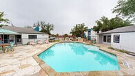 Located in Garland, TX, this outdoor community pool area offers a well-appointed retreat designed for relaxation and leisure. The centerpiece is a large, irregularly shaped swimming pool with clear turquoise water, framed by an expansive natural stone deck that provides ample space for sunbathing and socializing. Lounge chairs are positioned throughout the deck, accommodating residents seeking both sun and shade.

A covered pergola structure with a warm wood-toned ceiling anchors one side of the pool area, offering a shaded outdoor living space complete with seating — ideal for gathering or unwinding out of the sun. Mature trees are distributed around the perimeter, providing natural shade and a sense of established, lush greenery that softens the hardscape.

The pool area is enclosed by a wrought iron fence, ensuring security and privacy. A dedicated amenity building sits adjacent to the pool, finished in white brick and featuring signage, likely serving as a clubhouse, leasing office, or utility facility. An outdoor grill and dining area are also visible, adding functional entertaining space to the amenity zone.

The surrounding multi-story residential buildings feature a bold, modern color palette — deep charcoal and vivid blue exteriors — lending the overall environment a contemporary, updated aesthetic. The natural flagstone decking ties the space together with a cohesive, inviting character throughout.