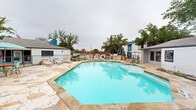 Located in Garland, TX, this outdoor community pool area offers a well-appointed retreat designed for relaxation and leisure. The centerpiece is a large, irregularly shaped swimming pool with clear turquoise water, framed by an expansive natural stone deck that provides ample space for sunbathing and socializing. Lounge chairs are positioned throughout the deck, accommodating residents seeking both sun and shade.

A covered pergola structure with a warm wood-toned ceiling anchors one side of the pool area, offering a shaded outdoor living space complete with seating — ideal for gathering or unwinding out of the sun. Mature trees are distributed around the perimeter, providing natural shade and a sense of established, lush greenery that softens the hardscape.

The pool area is enclosed by a wrought iron fence, ensuring security and privacy. A dedicated amenity building sits adjacent to the pool, finished in white brick and featuring signage, likely serving as a clubhouse, leasing office, or utility facility. An outdoor grill and dining area are also visible, adding functional entertaining space to the amenity zone.

The surrounding multi-story residential buildings feature a bold, modern color palette — deep charcoal and vivid blue exteriors — lending the overall environment a contemporary, updated aesthetic. The natural flagstone decking ties the space together with a cohesive, inviting character throughout.