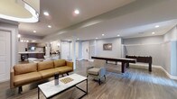The lounge at Summit at Mill Ridge is a well-appointed community space designed to encourage socializing and leisure. The open, single-floor layout is anchored by warm hardwood-style flooring that runs throughout, complementing the soft grey walls and crisp white trim for a clean, contemporary aesthetic.

The seating area features a rich caramel leather sofa paired with a upholstered accent chair, arranged around a marble-top coffee table — a thoughtful combination of comfort and modern style. A chessboard set on the table adds a welcoming, interactive touch.

The game zone includes a regulation shuffleboard table positioned along one wall, providing an entertaining focal point for residents and guests. Recessed lighting is distributed evenly across the ceiling, ensuring the entire space is well-lit and inviting throughout the day and evening.

Toward the back of the lounge, a wet bar area is visible, offering a convenient amenity for gatherings. Structural columns are seamlessly integrated into the design, while wall sconces add a warm, ambient glow. The overall atmosphere is relaxed yet refined, making it an ideal communal retreat.