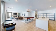 Located at 4101 William Turner Pkwy in Nashville, TN, this stylish single-floor community space offers a well-appointed, open-concept layout designed for both socializing and relaxation. The kitchen area is a standout feature, showcasing crisp white cabinetry, a striking black zellige tile backsplash, floating wood shelves with decorative accents, a large central island with seating, stainless steel appliances, and warm wood-look tile flooring that flows throughout the entire space. A pool table occupies the central gathering zone, adding a recreational focal point between the kitchen and lounge areas. The living room is anchored by a sleek linear fireplace set within a textured tile surround, with a wall-mounted flat-screen TV above. Richly upholstered seating, wood ottoman benches, and layered area rugs create an inviting atmosphere, while teal-toned accent walls add a sophisticated pop of color. A dedicated dining area features a large wood table surrounded by plush velvet chairs, ideal for group gatherings. Floor-to-ceiling sliding glass doors along the perimeter flood the space with natural light and provide direct views of and access to the outdoor pool deck, seamlessly blending indoor and outdoor living in a contemporary Nashville setting.