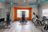 Located at 100 Stonegate Way in Middle Island, NY, this single-floor fitness center is designed to provide a well-equipped and energizing workout environment. The space features a bold, contemporary aesthetic defined by striking teal and orange accent colors that bring energy and personality to the room. Exposed teal ductwork runs across the vaulted ceiling, adding an industrial-chic element, while recessed lighting ensures the space is well illuminated throughout.

The flooring throughout is a light hardwood-style material that is both durable and visually clean. Large wall-mounted mirrors line one side of the room, creating a sense of expanded space and allowing users to monitor their form during workouts. Motivational fitness-themed typography decorates accent walls, reinforcing the active atmosphere.

The facility is outfitted with a comprehensive range of equipment, including treadmills, elliptical machines, stationary bikes, cable weight machines, a leg press, a dumbbell rack with a flat bench, and a multi-function cable training station. Natural light filters in through windows framed by the signature orange accent walls, contributing to an open and inviting feel. Wall-mounted televisions and a wall clock add practical amenities for users. Entry is provided through a distinctive orange-framed glass-paned door, completing the cohesive and thoughtfully designed space.