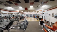 This fitness center offers a spacious, single-floor layout designed to accommodate a diverse range of workout routines. The facility is outfitted with an extensive selection of cardio machines, including ellipticals and rowing equipment, positioned throughout the open floor plan for easy accessibility. Strength training equipment and free weights are also well-represented, providing options for both beginner and advanced users.

The interior features a dropped tile ceiling with recessed fluorescent lighting that evenly illuminates the workout floor, ensuring a well-lit environment throughout. A bold orange and red accent stripe runs along the upper walls, adding a energetic visual element to the space. Floor-to-ceiling mirrors line the walls, enhancing the sense of depth and allowing users to monitor their form during exercise.

The flooring consists of durable, gym-appropriate material suited for high-traffic use. The open layout encourages efficient traffic flow between equipment stations. Security cameras are visible throughout, contributing to a safe and monitored environment. Overall, the fitness center presents a functional and organized space built to support active, health-focused use.