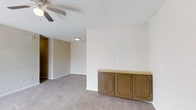 This single-floor space features a open living area finished with wall-to-wall neutral carpet, providing a soft and understated foundation throughout. The main room is equipped with a ceiling fan, offering both air circulation and overhead lighting, while a textured popcorn ceiling adds a classic touch to the interior. A notable built-in wooden cabinet with decorative panel detailing is positioned along one wall, providing practical storage and serving as a subtle design focal point. The warm brown tones of the cabinetry contrast with the bright white walls, creating a simple yet functional aesthetic. A hallway visible from the main living area leads further into the space, illuminated by a flush-mount ceiling light that ensures the passage remains well-lit. The walls are painted in a clean, neutral white throughout, lending the space a bright and airy feel. The overall layout is straightforward and functional, with an open flow between areas. The simplicity of the design allows for flexible furnishing and personalization, making it a practical and adaptable living environment.
