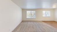 This single-floor, two-bedroom, one-bathroom unit at 1794 SW Fellows St in McMinnville, Oregon offers a efficiently arranged layout across a compact but functional footprint. The open-concept kitchen and living area serves as the heart of the unit, where light wood-tone cabinetry, white countertops, and a peninsula with an undermount sink and matte black faucet define the kitchen space. White appliances, including a range and refrigerator, complement the cabinetry, while luxury vinyl plank flooring runs throughout this area. The adjoining living room receives generous natural light through multiple windows with horizontal blinds, and features neutral carpet that continues into both bedrooms. Each bedroom is modestly sized, with the primary bedroom offering a slightly larger footprint, and both rooms include full-width sliding closets with six-panel doors. Windows in each bedroom provide outdoor views of surrounding greenery. The bathroom features vinyl plank flooring, a light wood vanity, and a full tub-shower combination with a chrome curtain rod. Cream-toned walls and clean white trim throughout lend the unit a bright, neutral aesthetic.